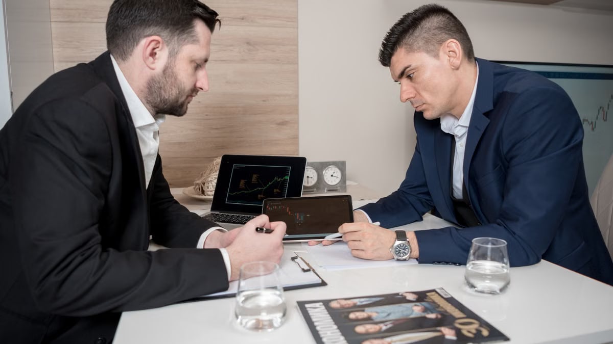 Two businessmen in suits discussing cryptocurrency trends using digital devices in an office setting.